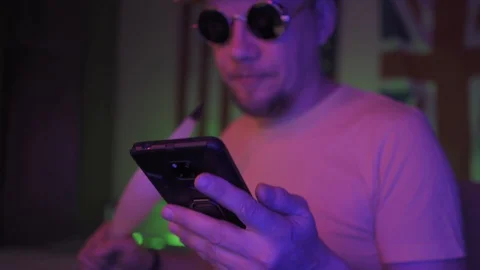 A man looking at the screen of a cell phone smoking hookah in neon lights Stock Footage 130043714