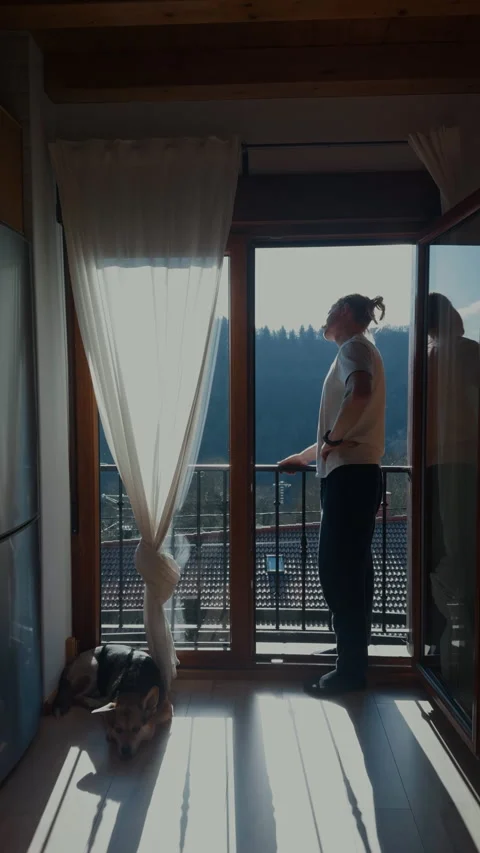 Man looking through balcony door enjoying morning view with dog at home Stock Footage 332558547