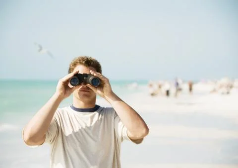 Man looking through binoculars on beach, facing camera Stock Photos