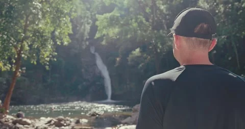 Man looking at tropical waterfall in lush green jungle 스톡 동영상 336298144