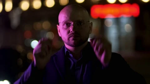 A man looks into the camera and adjusts glasses on the street at night 스톡 동영상 193061593