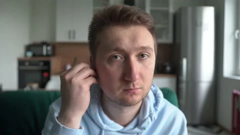 A man looks at the camera at home, inserts a wireless headset into his ears Stock Footage 154843257