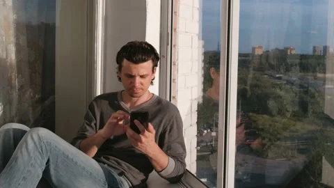 Man looks at mobile phone, types message sitting on the balcony at sunset. Video stock 102448927