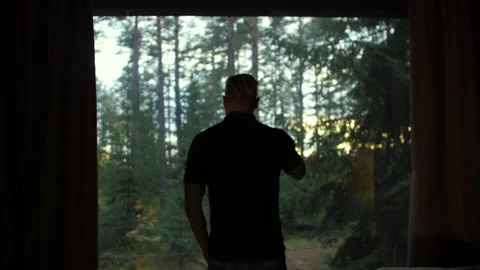 A man looks out the window in a forest house and drinks coffee. 스톡 동영상 184078291