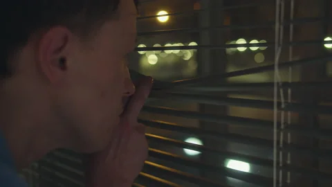 Man looks out the window through the blinds, the night city outside the window Stock Footage 158705437
