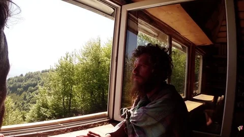 A man looks thoughtfully out the window at the mountains, in a wooden house Stock Footage 77785636