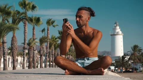 Man in lotus pose on waterfront, engrossed in smartphone video call with pa.. Stock Footage 344059210