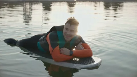 Man lying and smiling on a surfboard wearing wetsuit at warm golden sunset Stock-Footage 166545815