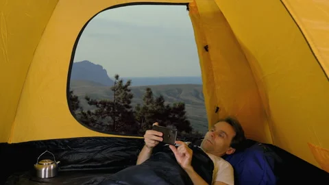 Man lying and using smartphone in open tent, scenic landscape in background Stock-Footage 161025495