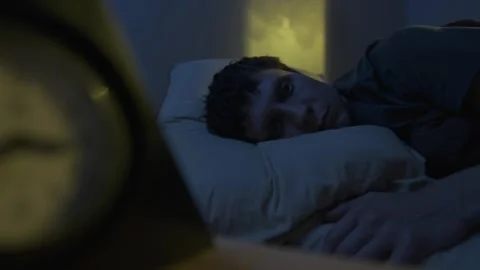 Man lying awake at night, blurry clock silhouette in foreground, warm yellow Vídeo Stock 320175423