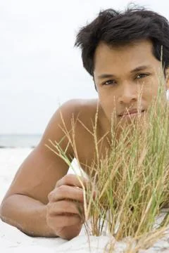 Man lying on the beach touching dune grass, looking at camera Stock Photos