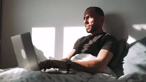 A man is lying on a bed and covered with a blanket working on a laptop A Stock Footage 154304125
