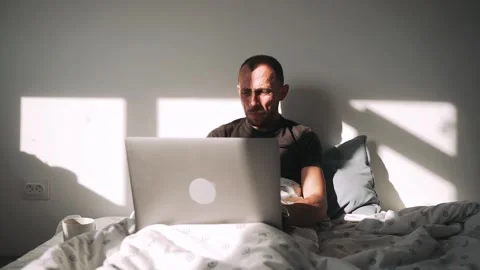 A man is lying on a bed and covered with a blanket working on a laptop A Stock Footage 154353301