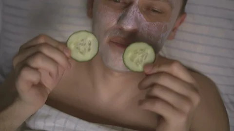 Man lying in bed cares for his face, sliced slices of cucumber on his eyes Stock Footage 130022304
