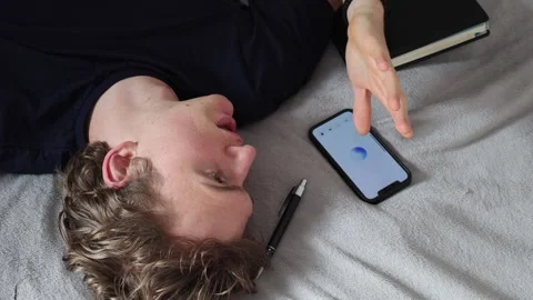 Man lying on bed gestures with hand while looking at smartphone screen experienc Stock Footage 314979759