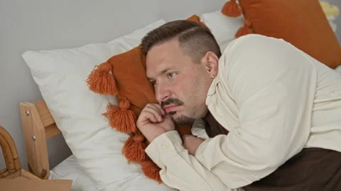 Man lying on bed with hand under chin by orange tassel pillow and white she.. 스톡 동영상 339213593