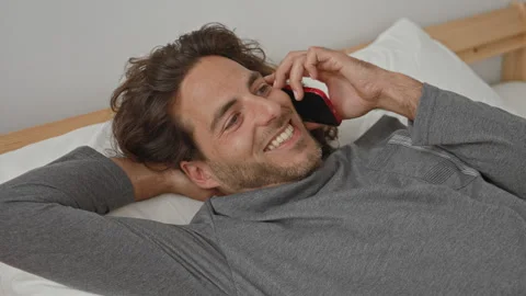 Man lying on bed holding smartphone to ear with hand behind head in a build.. Video stock 337377134