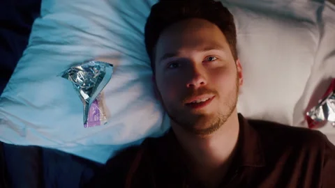 Man lying in bed at night with snack wrappers, looking thoughtful Vídeos de archivo 319441952