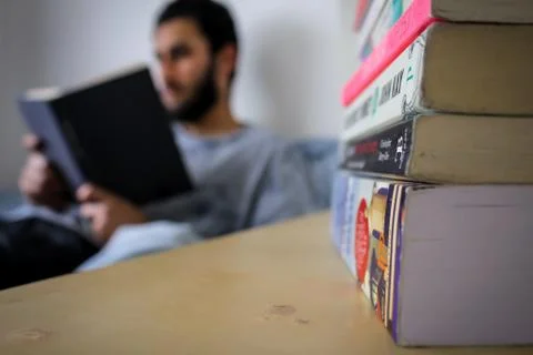 A man lying in bed reading a plane book with a stack of novels in focus Stock Photos