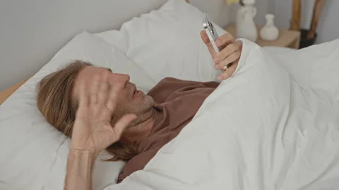 Man lying in bed scrolling a smartphone held above his face while touching .. Stock Footage 339194537