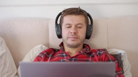 Man lying in bed wearing wireless headphones and looking at laptop screen Stock Footage 348761828