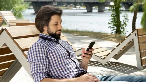 Man is lying on bench and holding phone. He is looking at it and typing message Stock-Footage 99740162