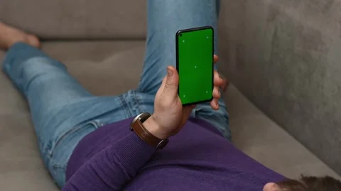 Man lying on couch holding phone with green Chroma Key screen mockup for web Stock Footage 259476519