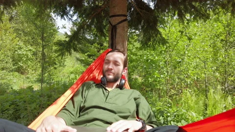 Man lying in hammock wearing wireless headphones openning top of laptop working  스톡 동영상 131899184