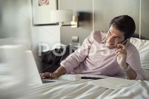 Man Lying On Hotel Bed, Talking On Cell Phone And Using Laptop Computer
