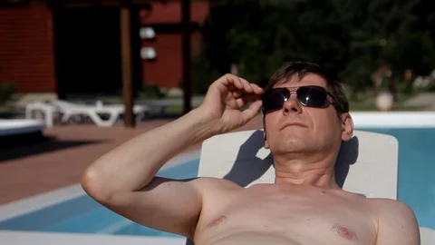 A man lying on a lounger by the pool raises sunglasses and squints Stock Footage 99935030
