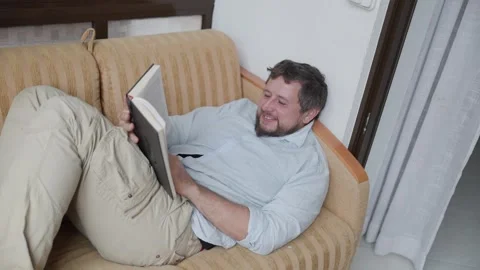 Man lying on sofa reading a book and showing different emotions Stock Footage 315743938