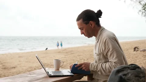 Man make notes to paper notebook, sit with laptop at working place on sea beach Stock Footage 147159767