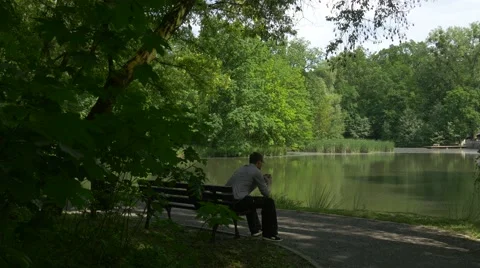 Man Makes a Call on Mobile Phone in Park Sitting Resting on a Bench by the Lake Stock Footage 63810752