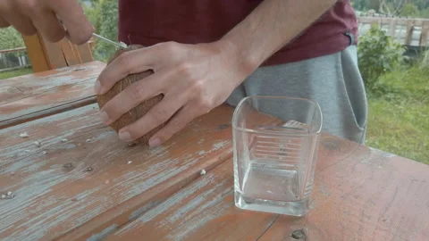 Man makes a hole in a coconut using knife, opens and pours coconut water into  Stockbeeldmateriaal 132701323