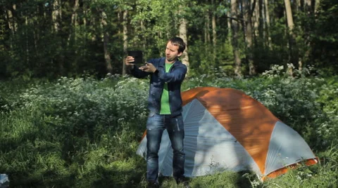 A man makes a video call on the tablet. Nearby stands a tent in the forest Stock Footage 64328454