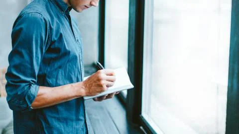 Man making notes in a notebook, standing near the window. Stock Photos
