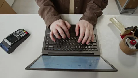 Man Managing Online Store Orders on Laptop at Counter with Payment Terminal Stock Footage 342589497