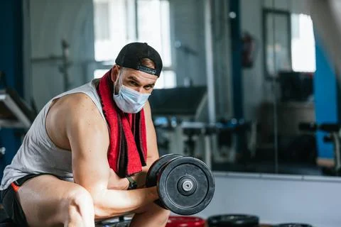 Man with mask doing biceps exercises with dumbbells in the gym. sitting on a  Stock Photos