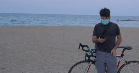 Man with mask looking at the smartphone on the beach with his bike Stock Footage 130944054