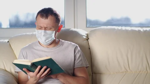 Man in mask reads a book while sitting on a sofa on a sunny day, moving camera Stock Footage 128311062