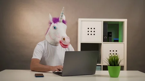The man in the mask of a unicorn sitting at a table working on laptop 動画素材 128998302