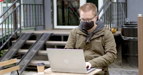 A man in a mask works on a laptop while sitting in a cafe on the street.  Stock Footage 142883773