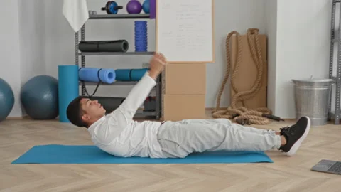 Man on mat doing situp with arms raised holding red ball, wearing white shi.. Stock Footage 339252107