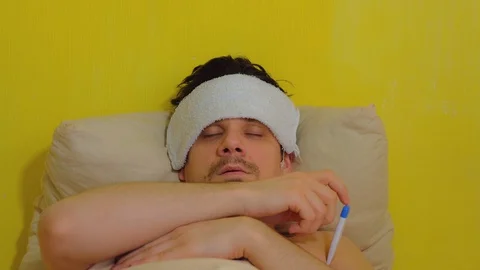 Man measures body temperature using thermometer and puts wet towel on forehead. Video stock 128959574