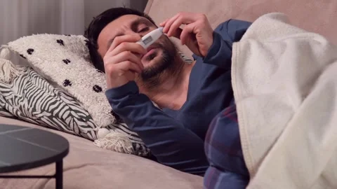 A man measures his body temperature with a thermometer while lying on a sofa Stock-Footage 323504449