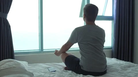 A man meditates on bed while looking out the window. Back view Video stock 130433333
