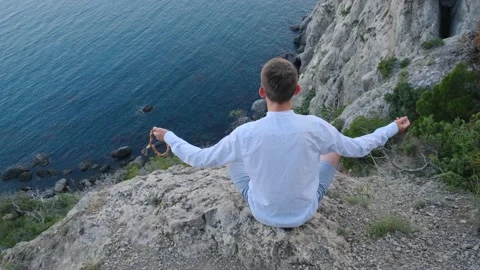 A man is meditating while sitting on the edge of a cliff over a pasture Stock Footage 159350916