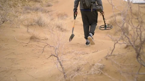 A man with a metal detector and a shovel walks through a sandy desert area Stock Footage 228100053