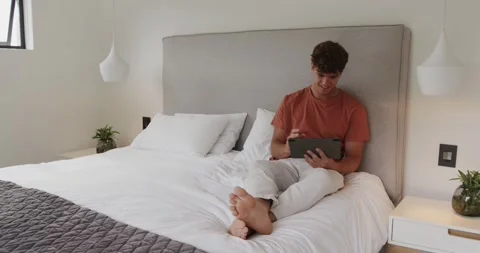 Man mid-20s settling cross-legged on bed tapping tablet to explore content and Stock Footage 316478667