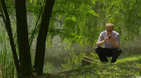 Man With Mobile Phone Calling Squatting at Lake Bank in Park Holding a Stock Footage 63810259